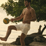 Man kicking a soccer ball on a beach with trees and water in the background.