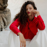 Woman in a red blouse and white pants sitting against a stone wall.