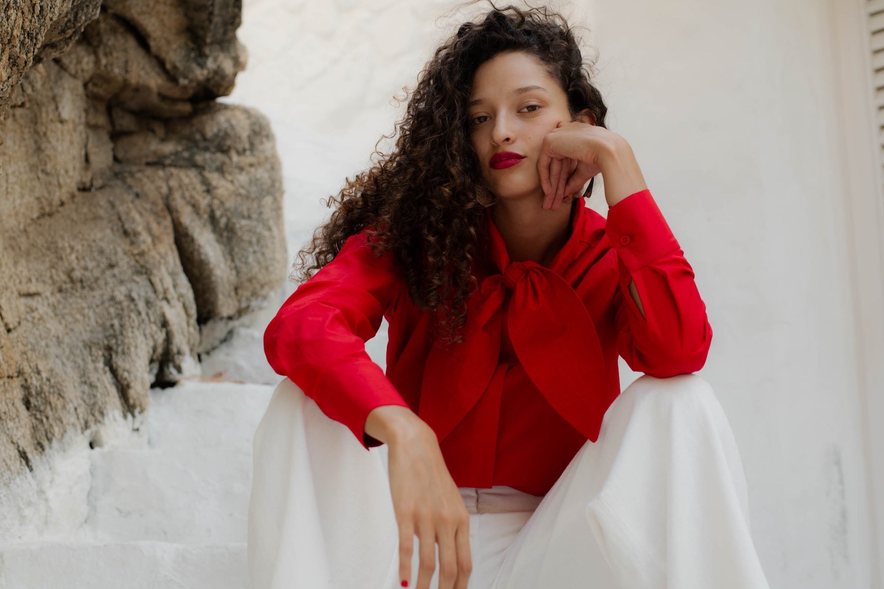 Woman in a red blouse and white pants sitting against a stone wall.