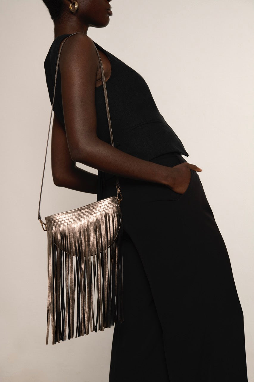 Person wearing a black outfit with a metallic gold fringe handbag on a plain background.