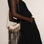 Person wearing a black outfit with a metallic gold fringe handbag on a plain background.