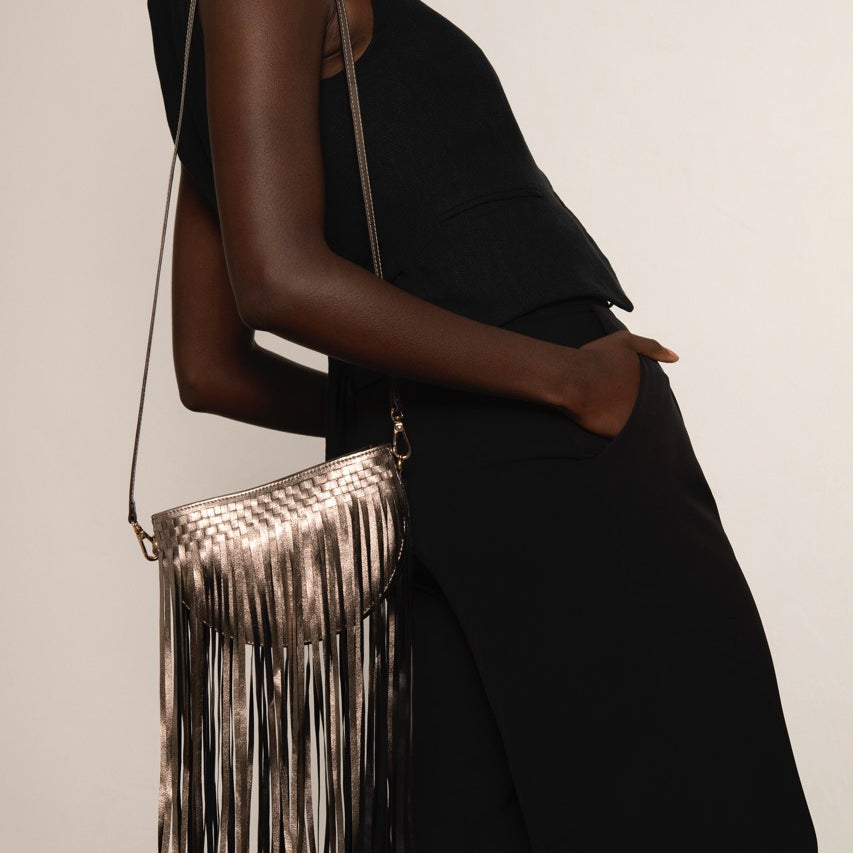 Person wearing a black outfit with a metallic gold fringe handbag on a plain background.