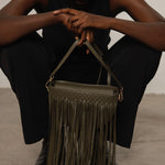 Person holding a green handbag with fringe details on a plain background.