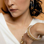 Close-up of a person wearing gold jewelry including earrings, rings, and a bracelet on a neutral background.