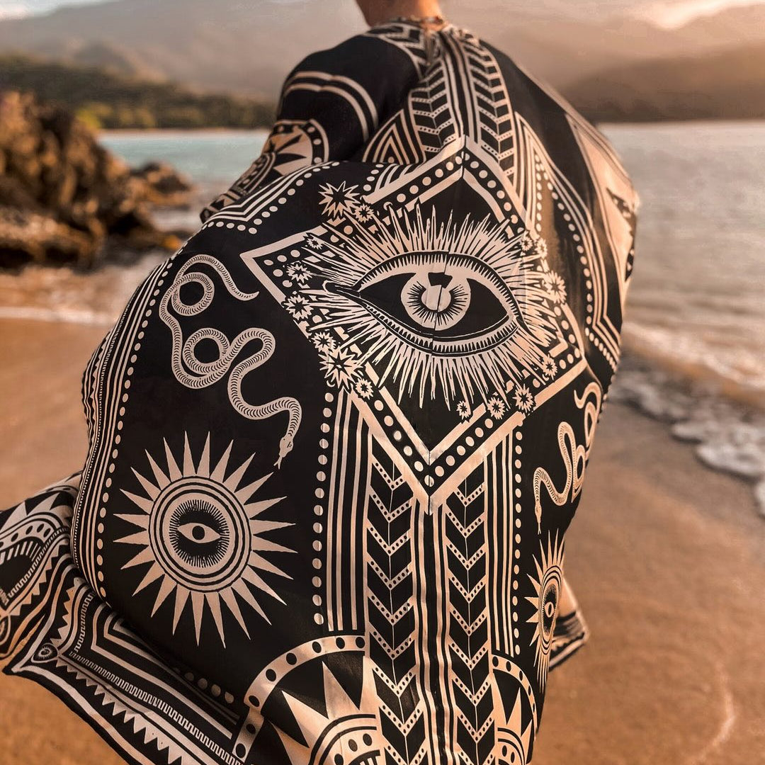 A person standing on a beach with their back to the camera, wrapped in a black and cream bohemian patterned kimono featuring eye and geometric designs.