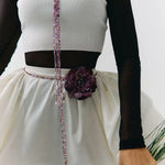 A close-up of a person wearing a white ribbed sleeveless top with black sheer long sleeves, a dark brown waistband, and a white skirt with a large dark purple flower at the waist. A beaded purple and pink lanyard hangs down the front, and the person is holding green stems against a plain light background.