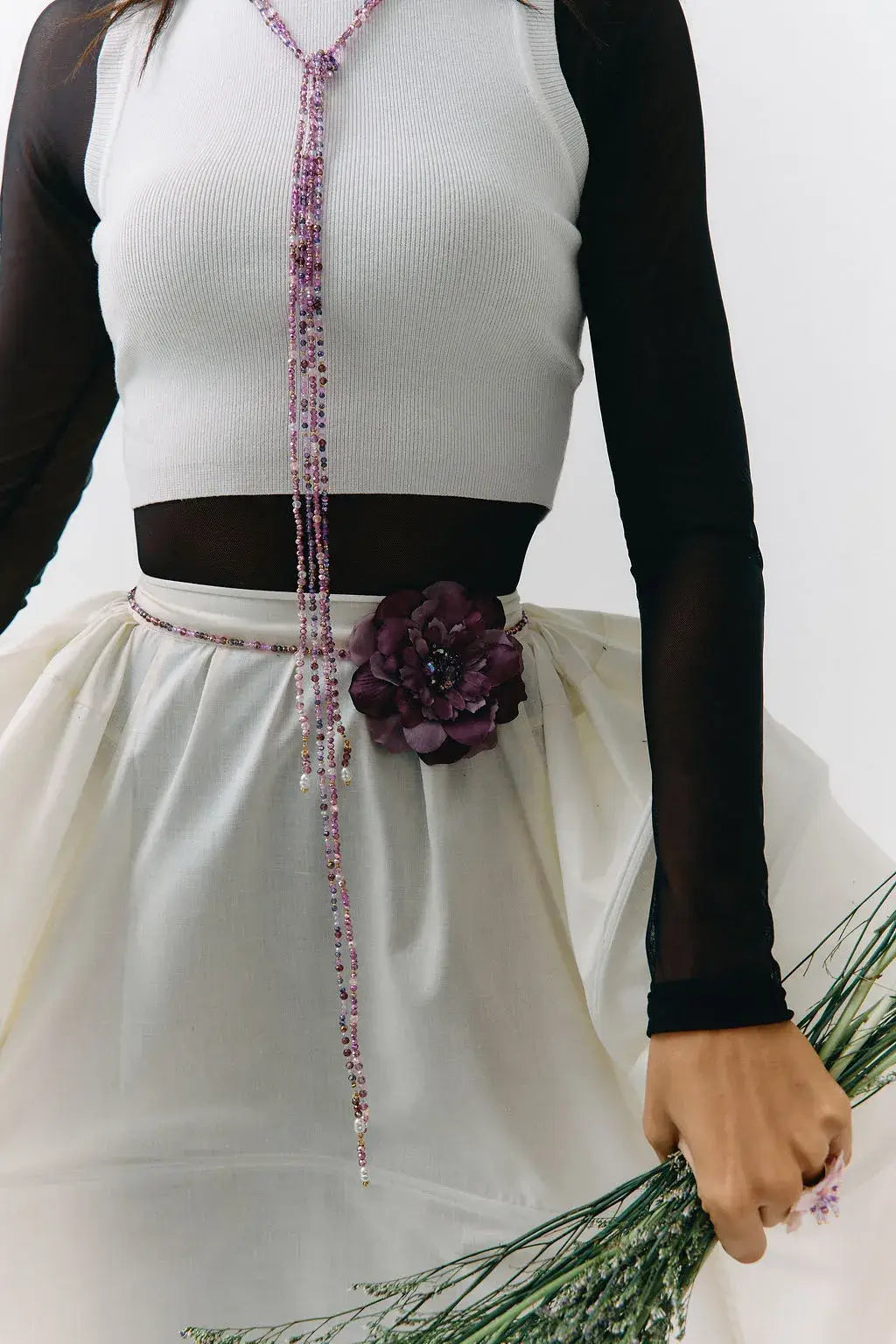 A close-up of a person wearing a white ribbed sleeveless top with black sheer long sleeves, a dark brown waistband, and a white skirt with a large dark purple flower at the waist. A beaded purple and pink lanyard hangs down the front, and the person is holding green stems against a plain light background.