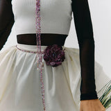 A close-up of a person wearing a white ribbed sleeveless top with black sheer long sleeves, a dark brown waistband, and a white skirt with a large dark purple flower at the waist. A beaded purple and pink lanyard hangs down the front, and the person is holding green stems against a plain light background.
