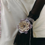 A close-up of a person wearing a white blouse and sheer black long gloves, with a decorative fabric flower accessory attached to one glove and a beaded bracelet on the wrist.
