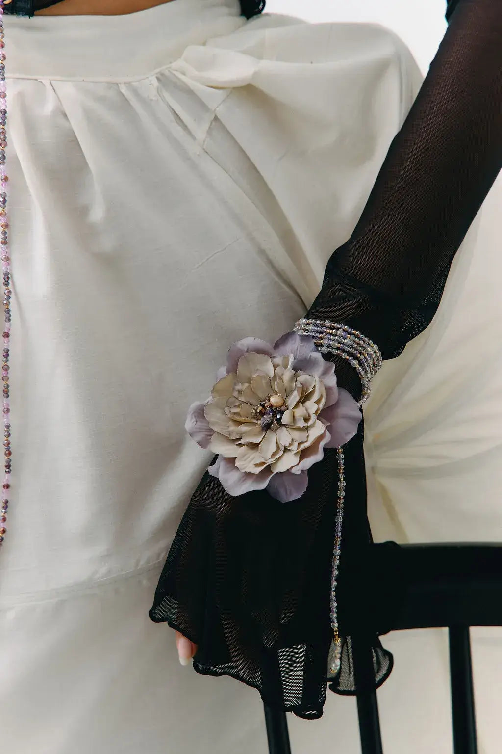 A close-up of a person wearing a white blouse and sheer black long gloves, with a decorative fabric flower accessory attached to one glove and a beaded bracelet on the wrist.