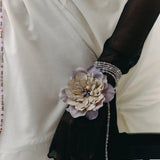 A close-up of a person wearing a white blouse and sheer black long gloves, with a decorative fabric flower accessory attached to one glove and a beaded bracelet on the wrist.