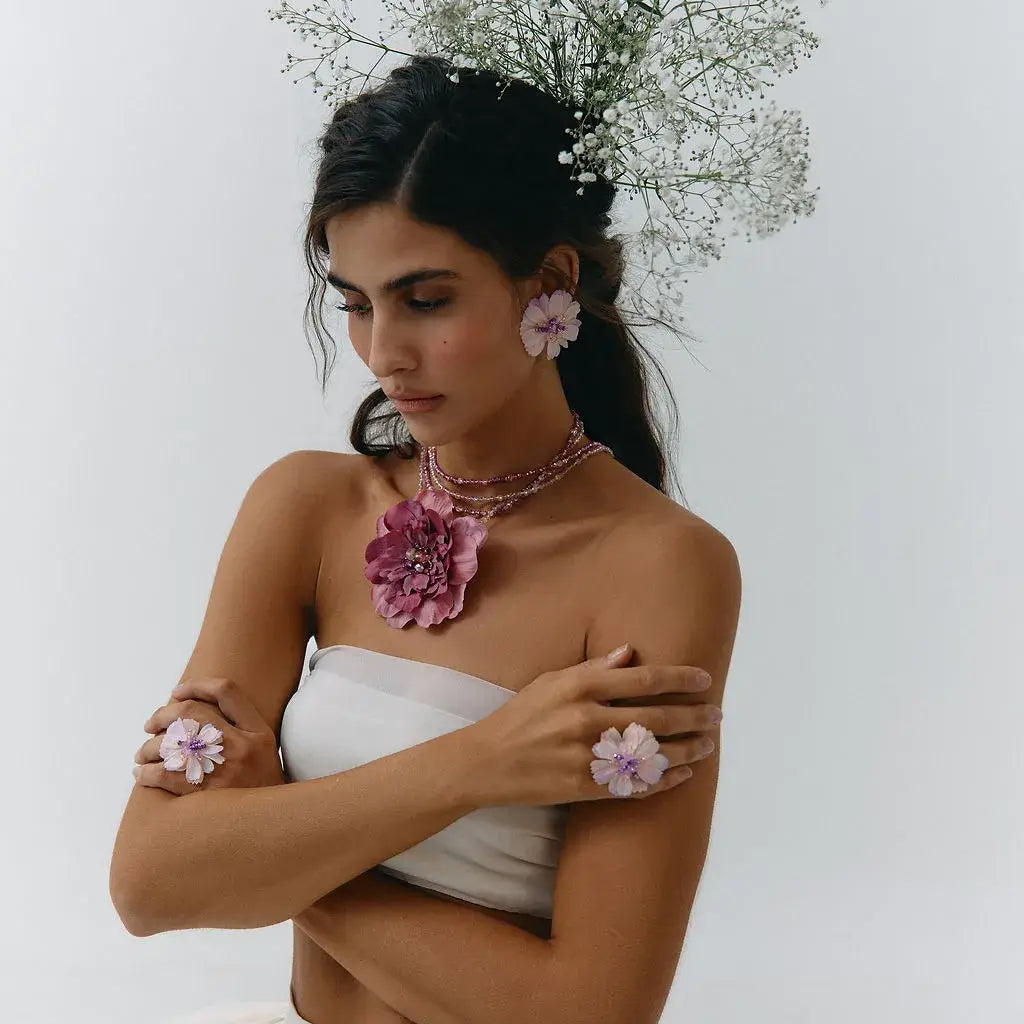 A woman in a white strapless dress poses with a floral headpiece, pink flower earrings, and pink flower bracelets against a plain white background.