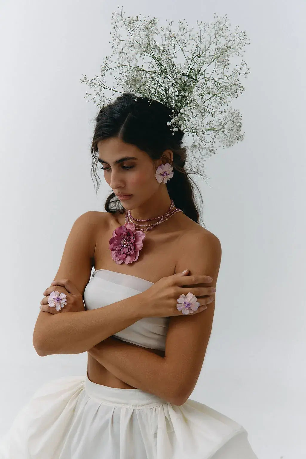 A woman in a white strapless dress poses with a floral headpiece, pink flower earrings, and pink flower bracelets against a plain white background.
