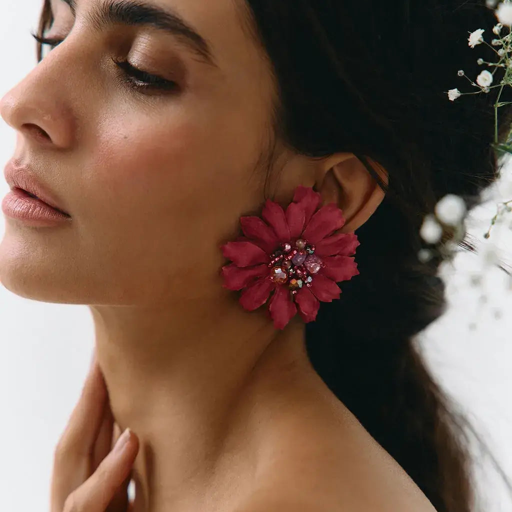 A close-up side profile of a woman wearing a large, vibrant magenta-pink flower-shaped earring with a jeweled center, set against a soft, light background.