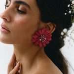 A close-up side profile of a woman wearing a large, vibrant magenta-pink flower-shaped earring with a jeweled center, set against a soft, light background.