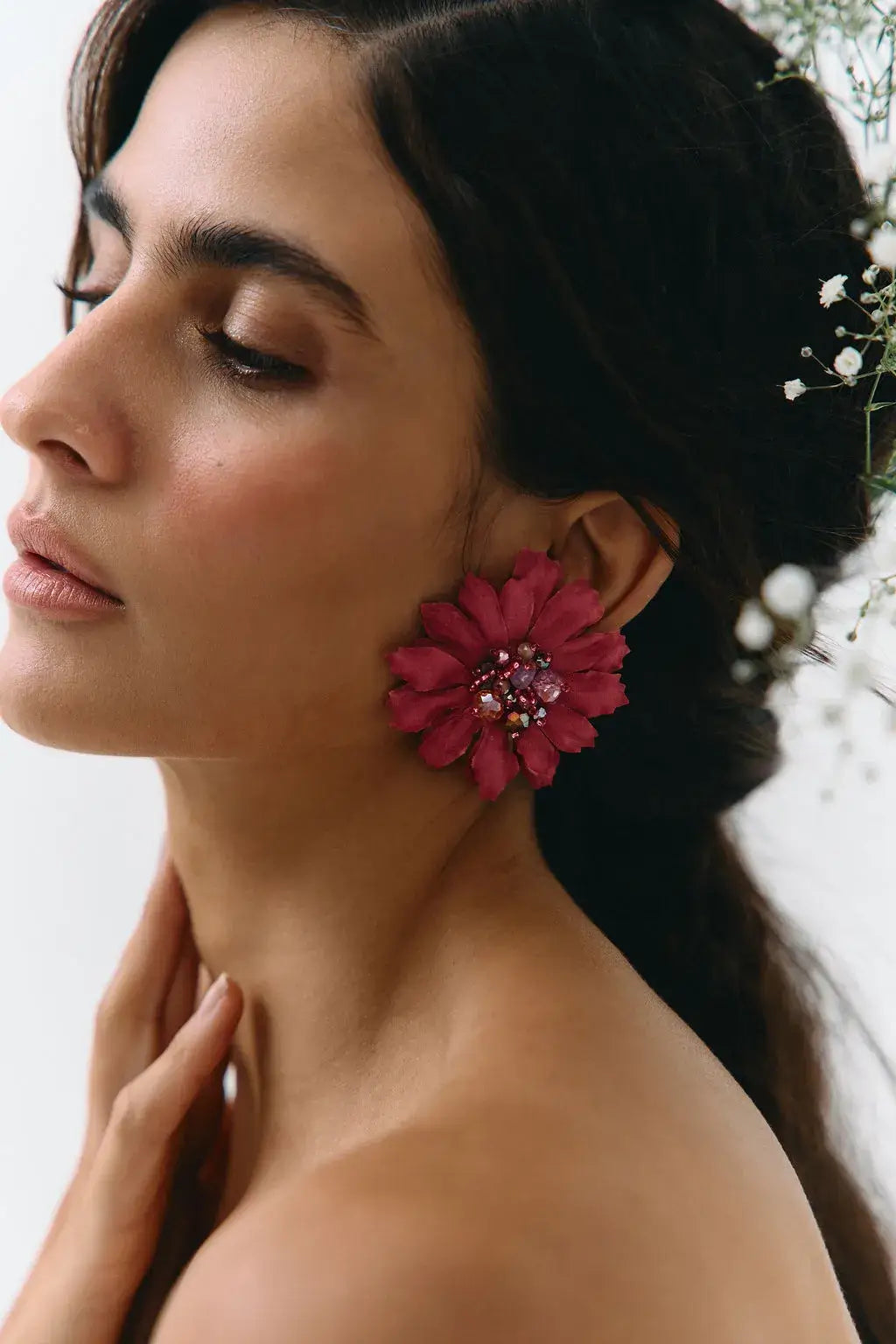 A close-up side profile of a woman wearing a large, vibrant magenta-pink flower-shaped earring with a jeweled center, set against a soft, light background.