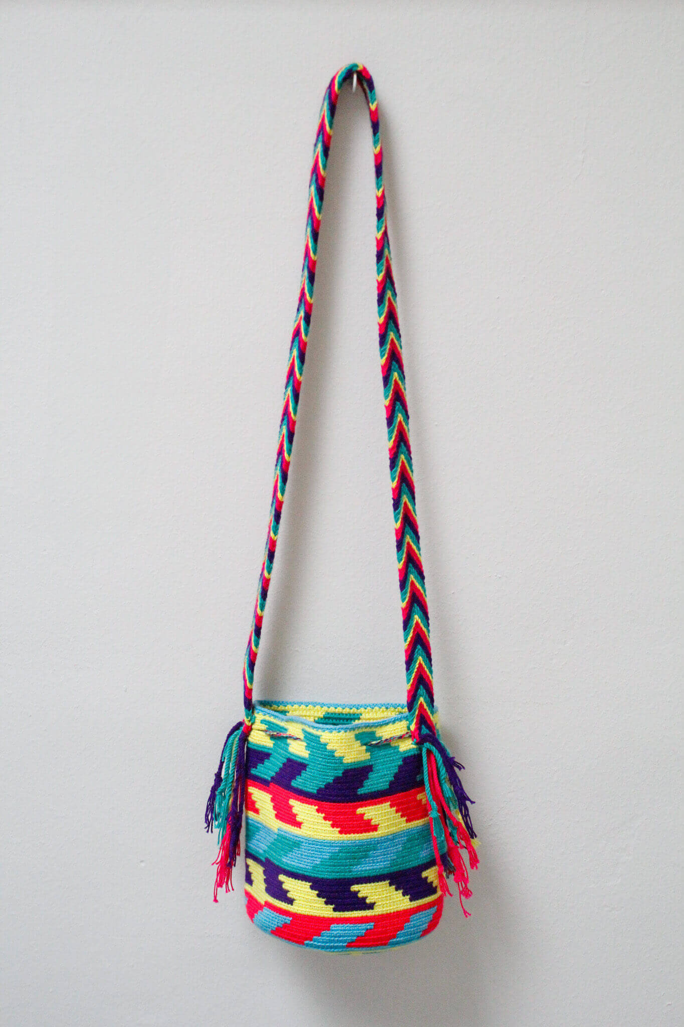 A small handmade Wayuu crossbody bag with a geometric pattern, featuring a wide woven strap and a flat tassel closure.