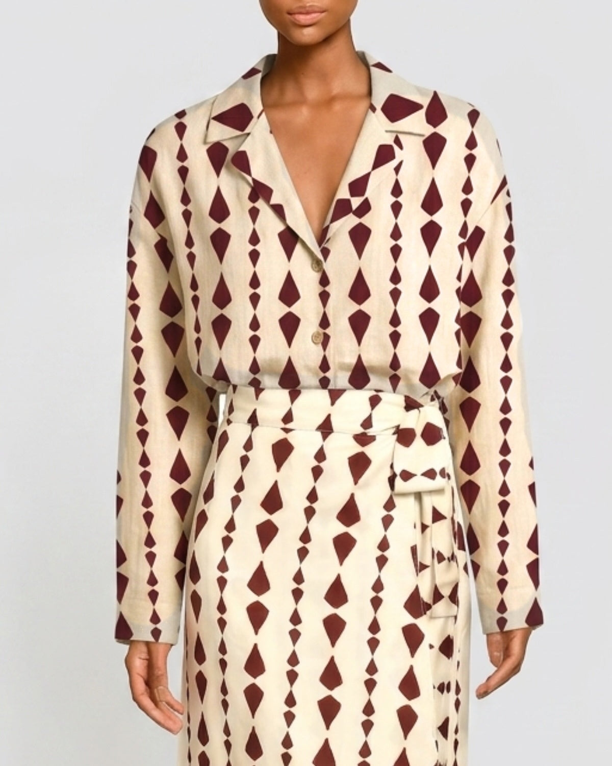 A model wearing a long-sleeve collared shirt dress with a cream base and a dark burgundy geometric diamond pattern, featuring a matching tie belt.