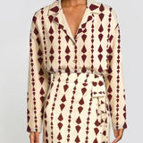 A model wearing a long-sleeve collared shirt dress with a cream base and a dark burgundy geometric diamond pattern, featuring a matching tie belt.