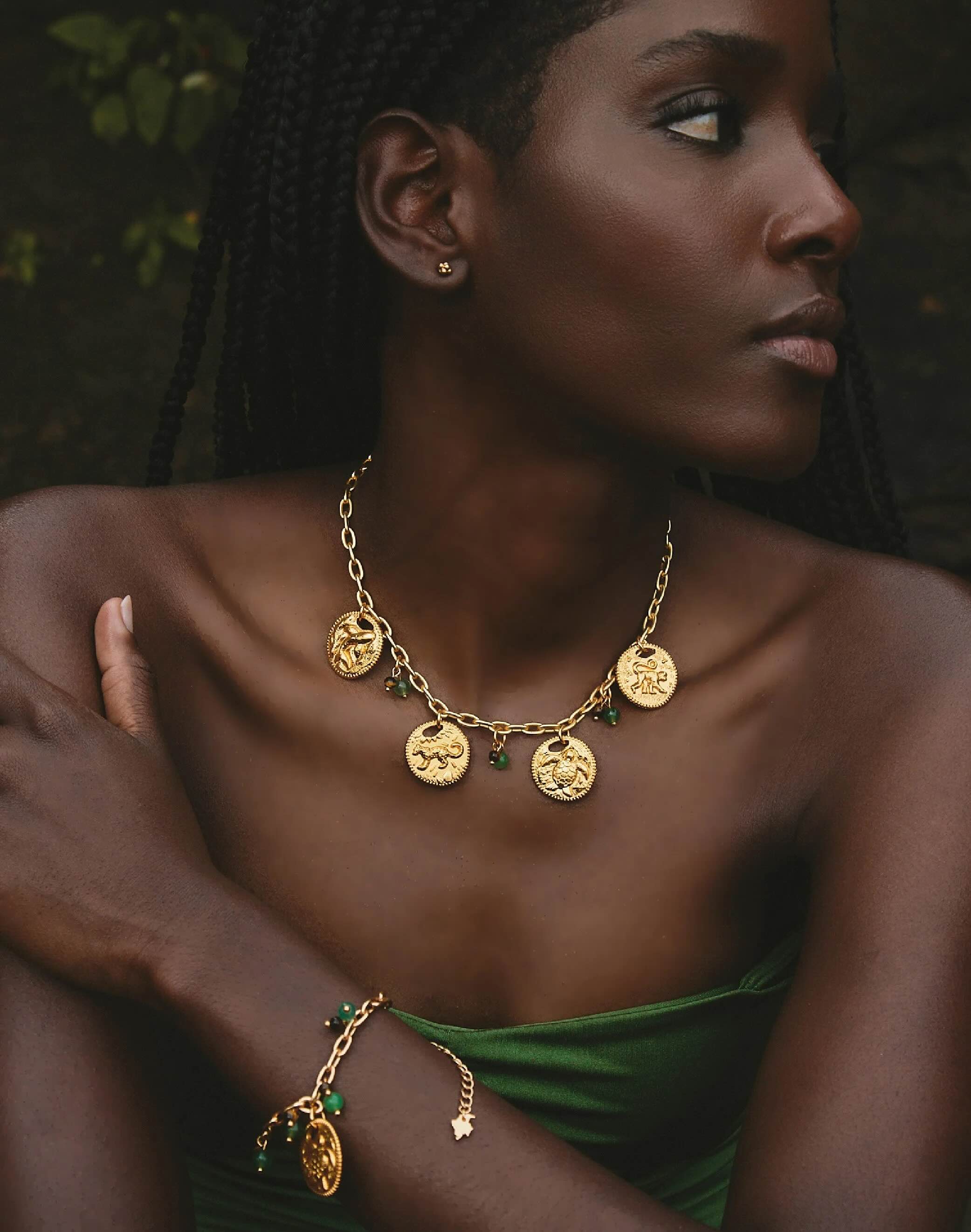 Woman wearing a gold necklace and bracelet with green accents against a dark background