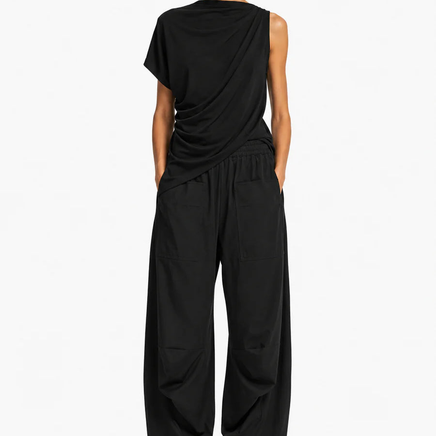 A model wearing a black draped asymmetrical top and matching wide-leg trousers, styled with black loafers against a plain white studio background.