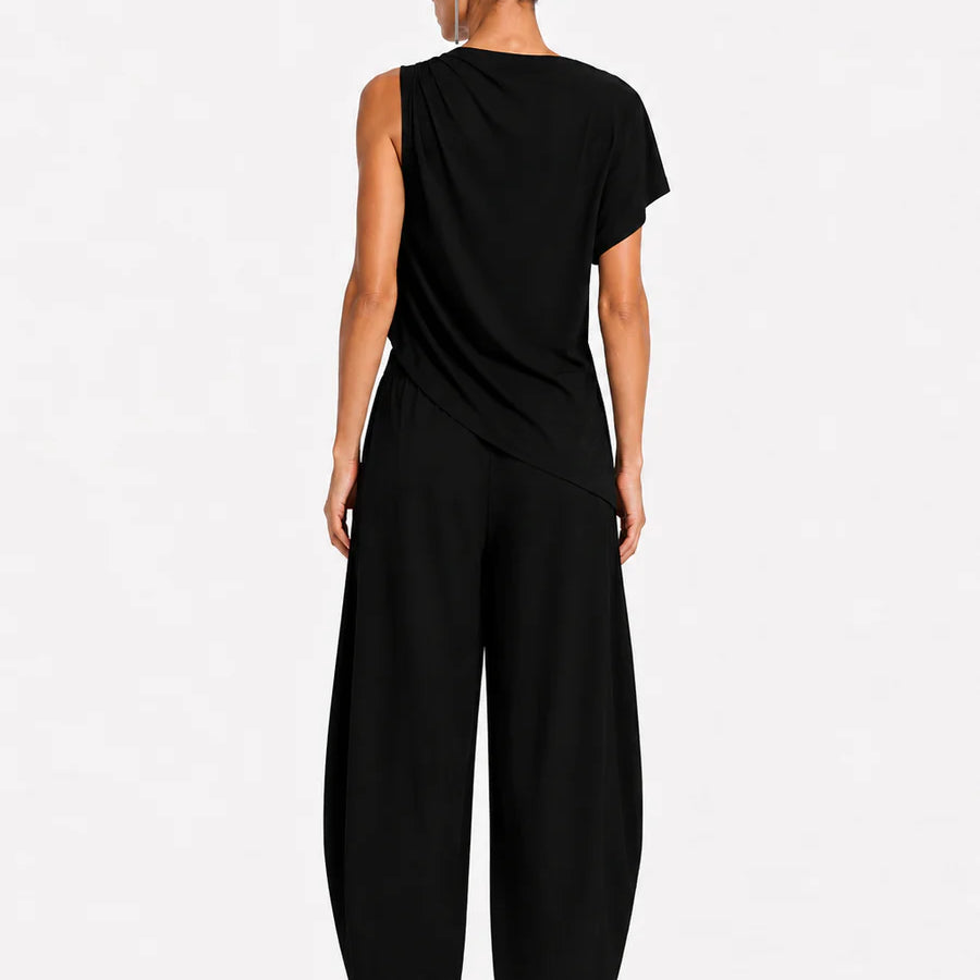 A model viewed from the back wearing a black draped sleeveless top and matching wide-leg pants, standing against a plain light gray studio background.