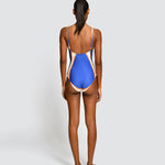 A woman shown from the back wearing a one-piece swimsuit with a royal blue body and beige side panels, standing against a light gray studio background.