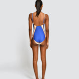 A woman shown from the back wearing a one-piece swimsuit with a royal blue body and beige side panels, standing against a light gray studio background.