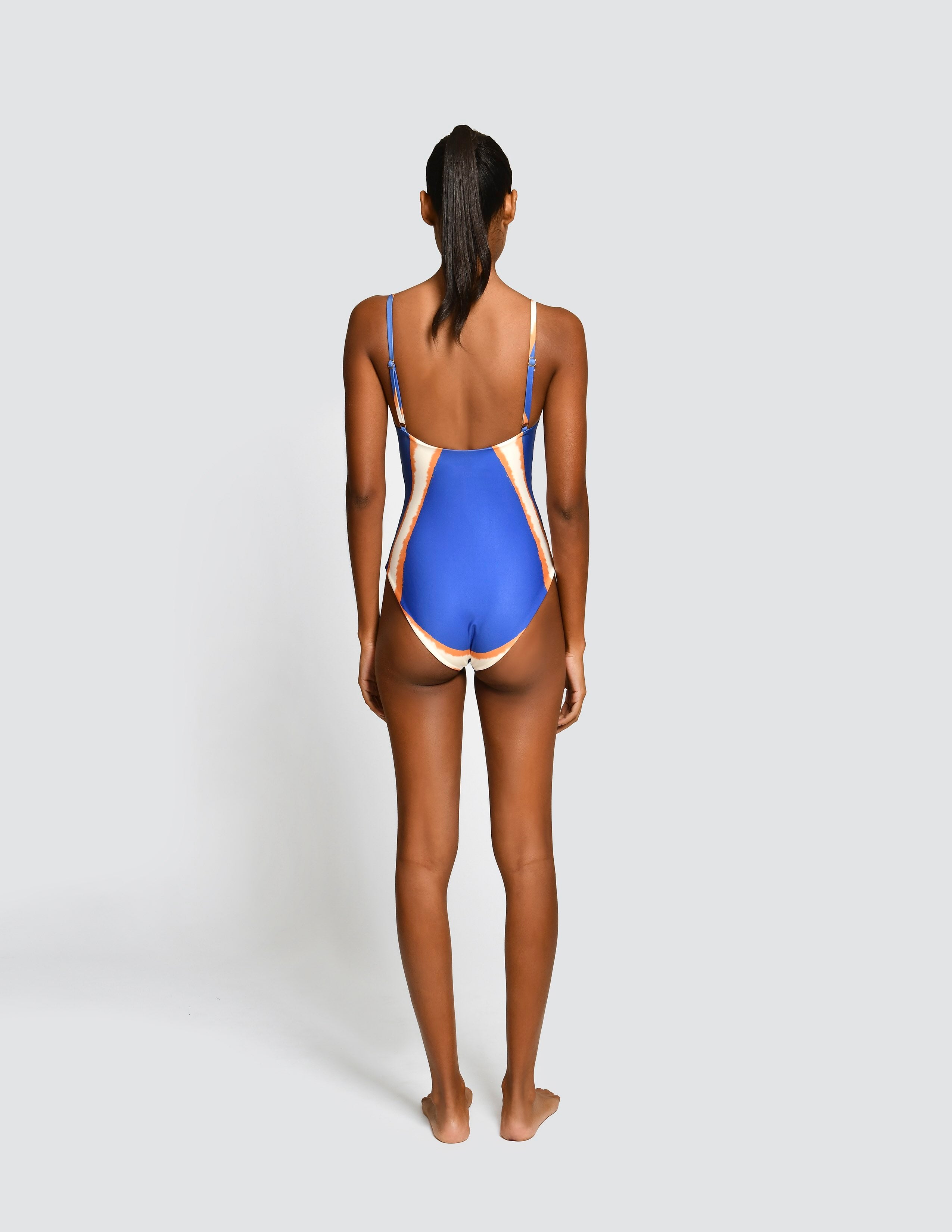 A woman shown from the back wearing a one-piece swimsuit with a royal blue body and beige side panels, standing against a light gray studio background.