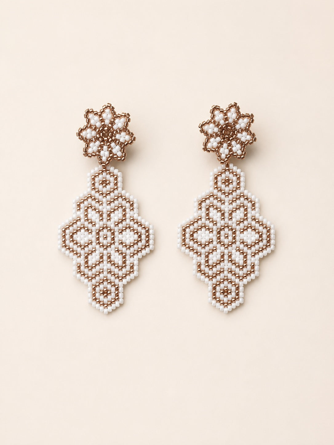 A pair of handmade beaded dangle earrings featuring intricate geometric and floral patterns in white and light brown seed beads, displayed on a solid light beige background.