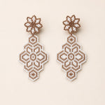 A pair of handmade beaded dangle earrings featuring intricate geometric and floral patterns in white and light brown seed beads, displayed on a solid light beige background.