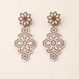 A pair of handmade beaded dangle earrings featuring intricate geometric and floral patterns in white and light brown seed beads, displayed on a solid light beige background.