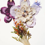 A hand-painted flower bouquet in a paintbrush handle, featuring a large lavender and cream flower with pearl-like accents and dried orange-brown filler, hanging from a string with wooden clothespins against a white background.
