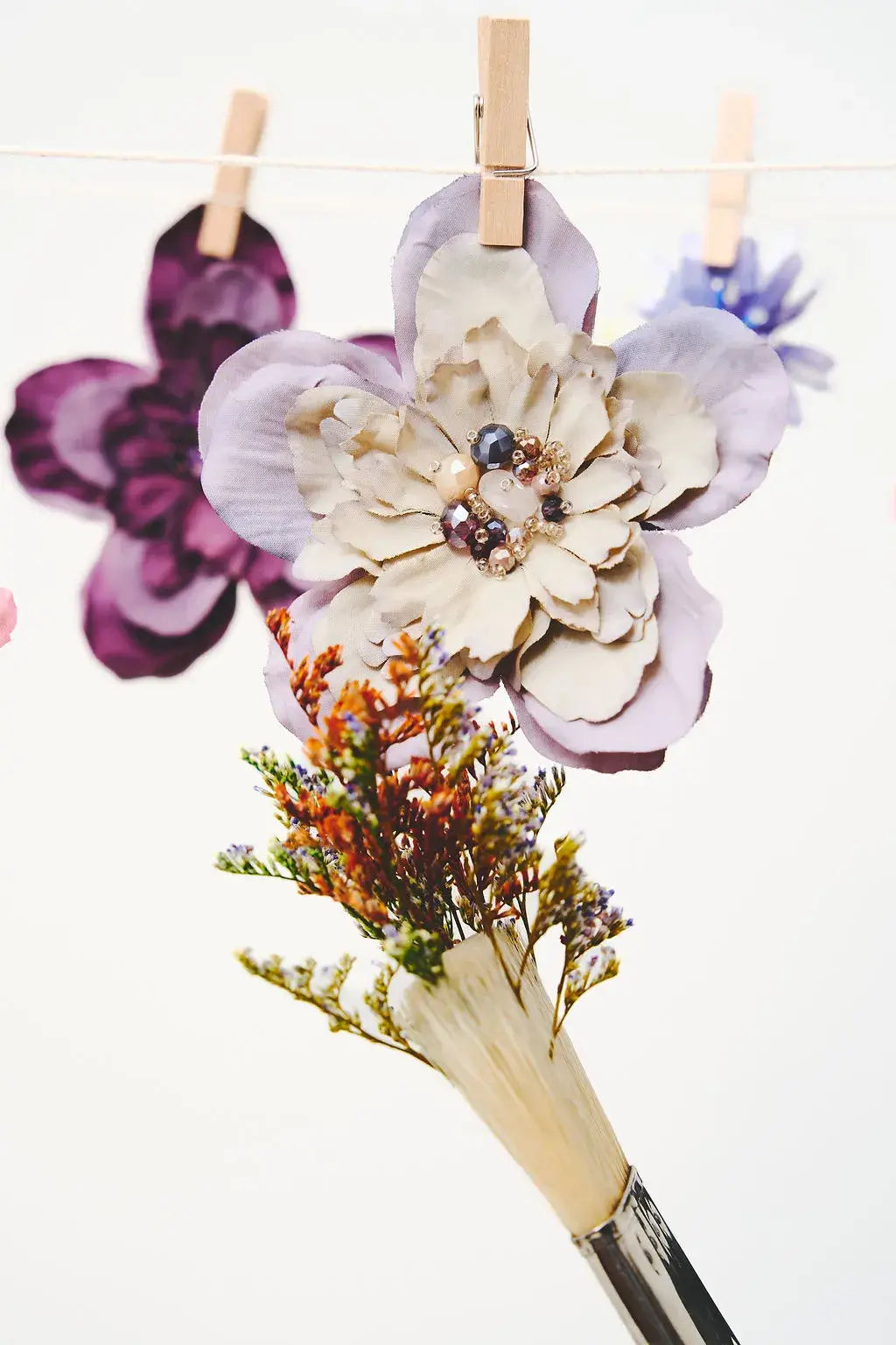 A hand-painted flower bouquet in a paintbrush handle, featuring a large lavender and cream flower with pearl-like accents and dried orange-brown filler, hanging from a string with wooden clothespins against a white background.
