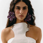A close-up portrait of a model wearing a white textured halter-neck top with gold chain accents and large purple flower hair clips against a plain white background.