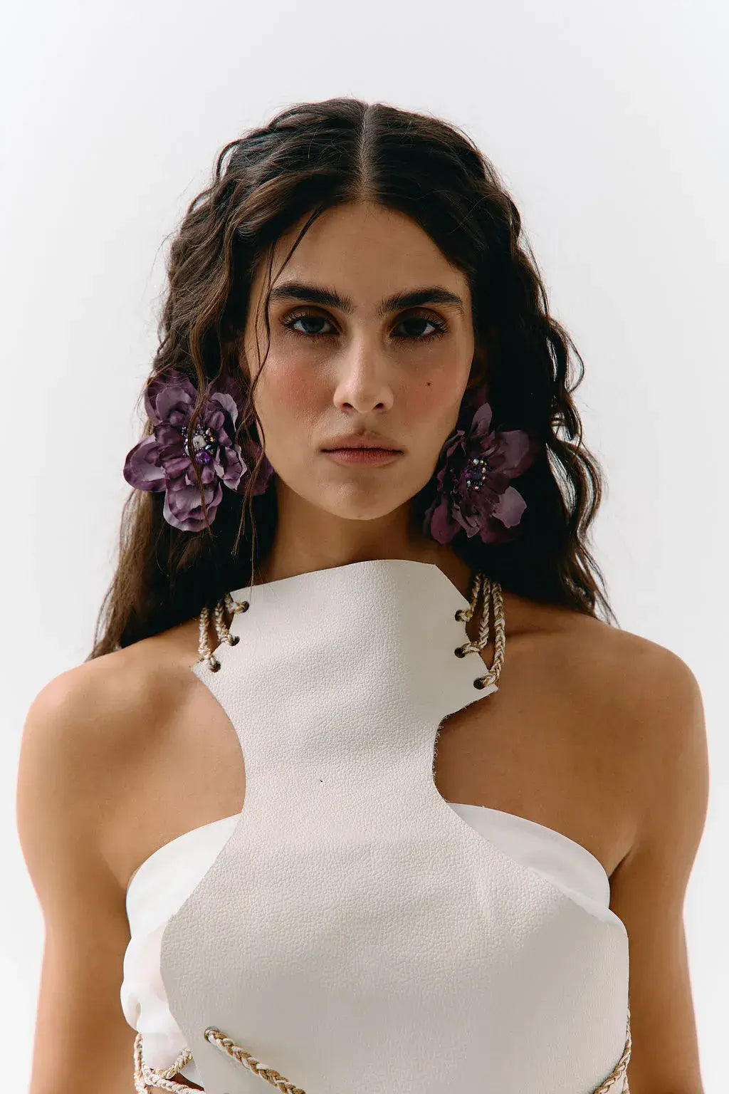 A close-up portrait of a model wearing a white textured halter-neck top with gold chain accents and large purple flower hair clips against a plain white background.
