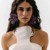 A close-up portrait of a model wearing a white textured halter-neck top with gold chain accents and large purple flower hair clips against a plain white background.