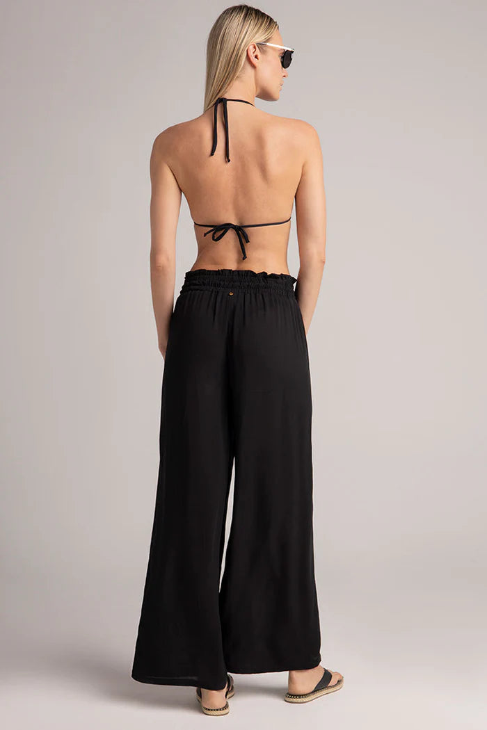 Woman wearing a black halter bikini top and wide-leg pants on a plain background.