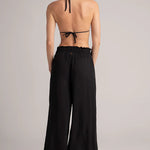 Woman wearing a black halter bikini top and wide-leg pants on a plain background.