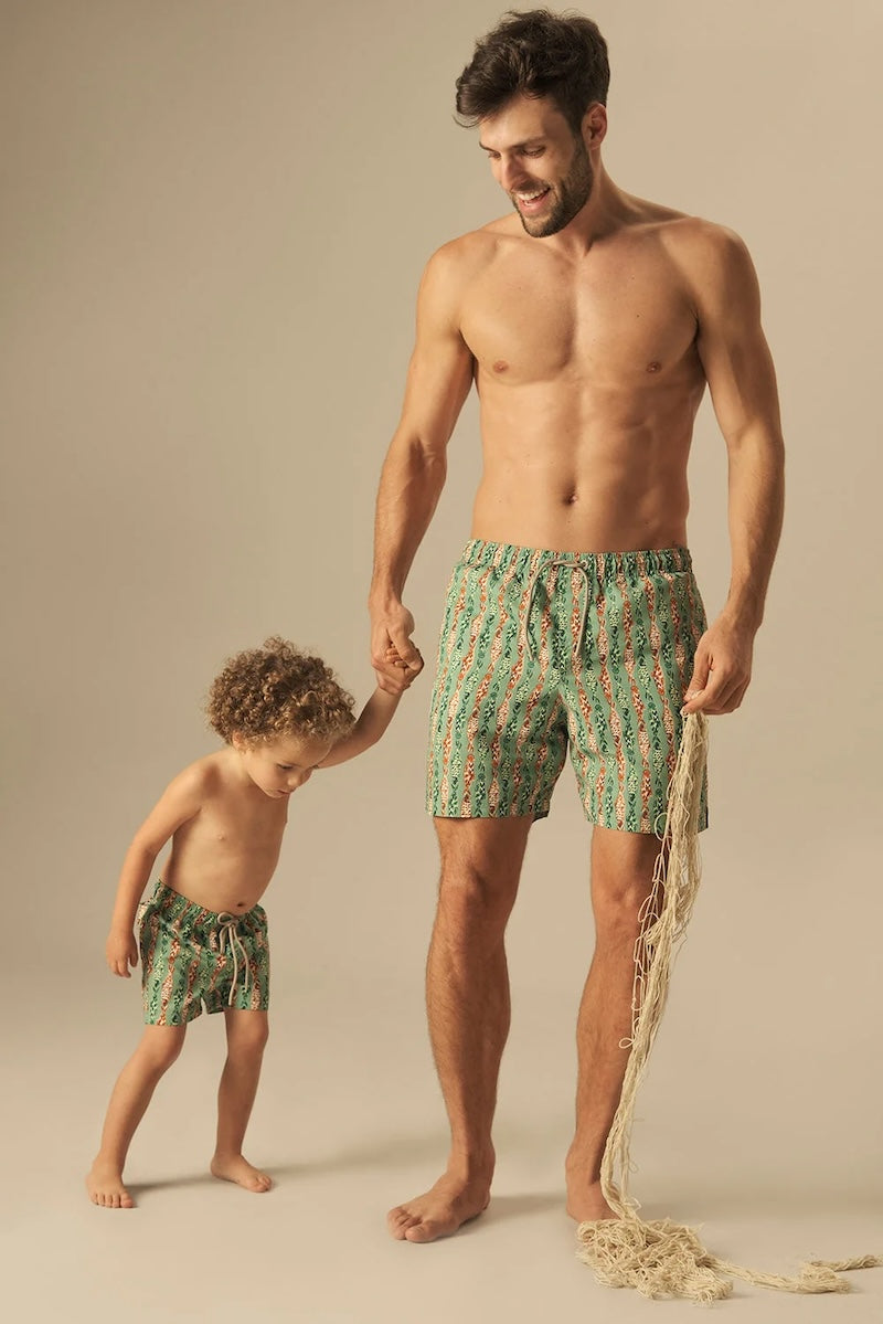 Man and child wearing matching green and orange fish pattered swim trunks on a beige background. 