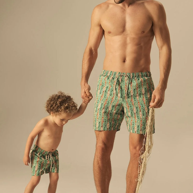 Man and child wearing matching green and orange fish pattered swim trunks on a beige background. 