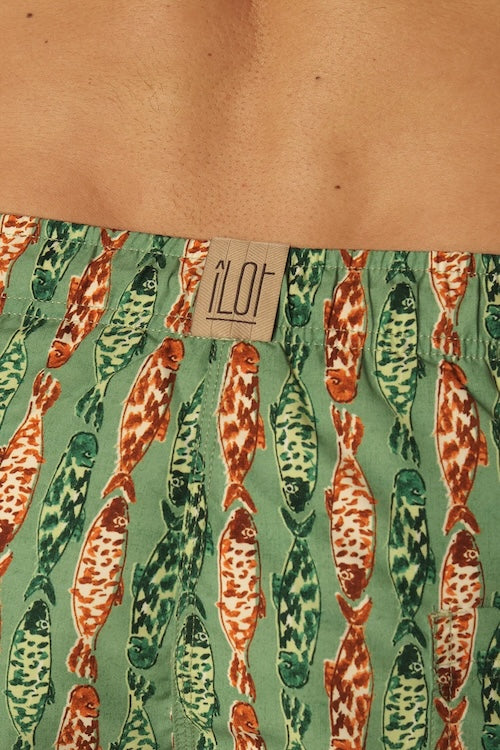 Close up of green and orange fish-patterned swim shorts with a brand label on a beige background.