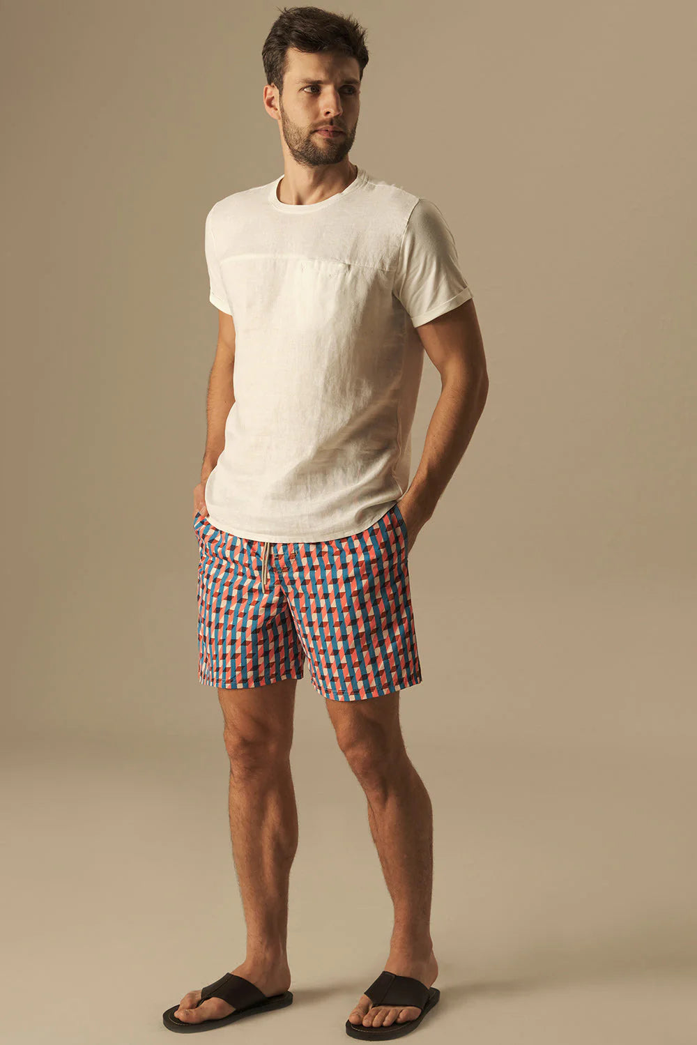 Man wearing a white t-shirt and checkered swim shorts on a beige background.