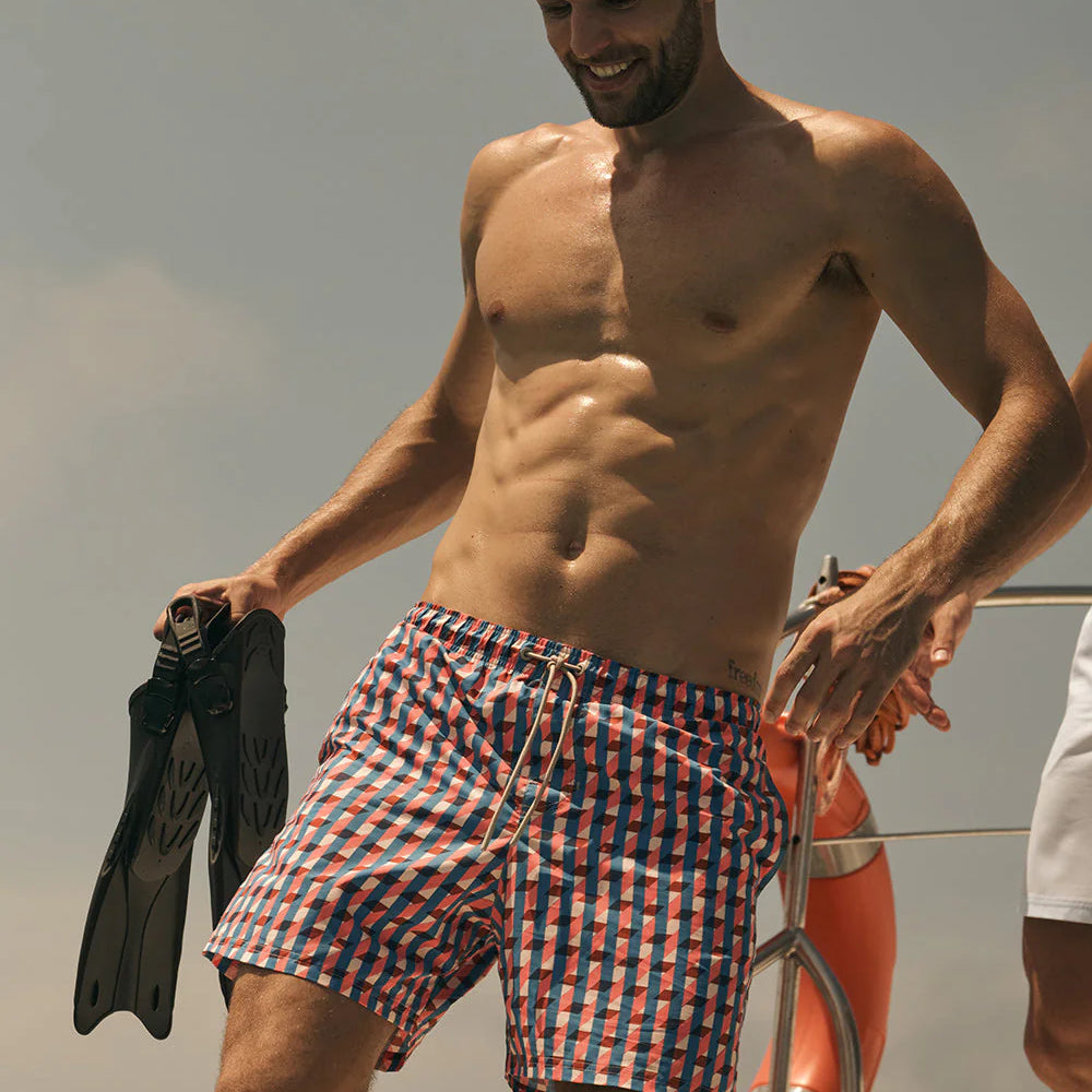 Man on a boat wearing checkered swim shorts and a diving mask, holding flippers.