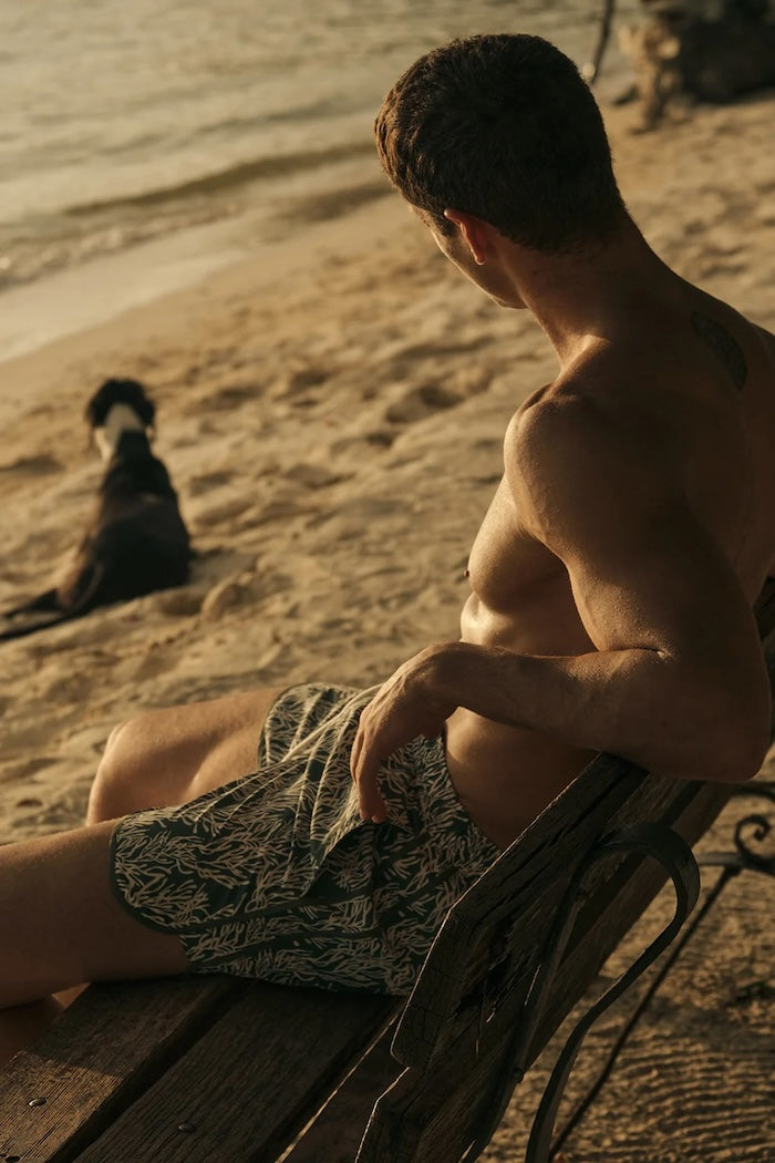 Man sitting on a wooden bench by the beach with a dog in the background wearing green swim trunks. 