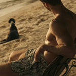 Man sitting on a wooden bench by the beach with a dog in the background wearing green swim trunks. 