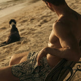 Man sitting on a wooden bench by the beach with a dog in the background wearing green swim trunks. 