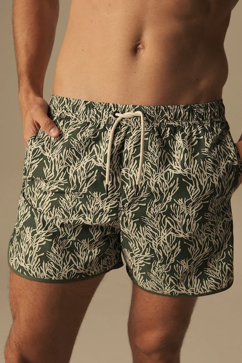 Green and white patterned swim shorts worn by a person on a neutral background.