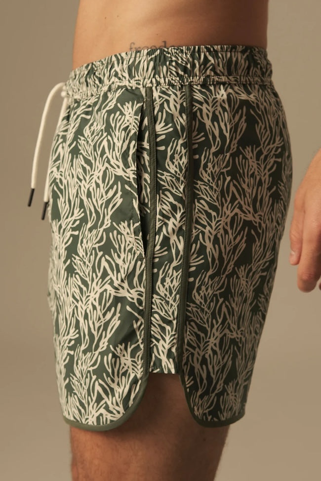 Green and white leaf patterned swim trunks worn by a person on a neutral background.