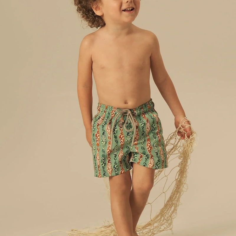 Child wearing green and orange fish patterned shorts standing on a beige background.